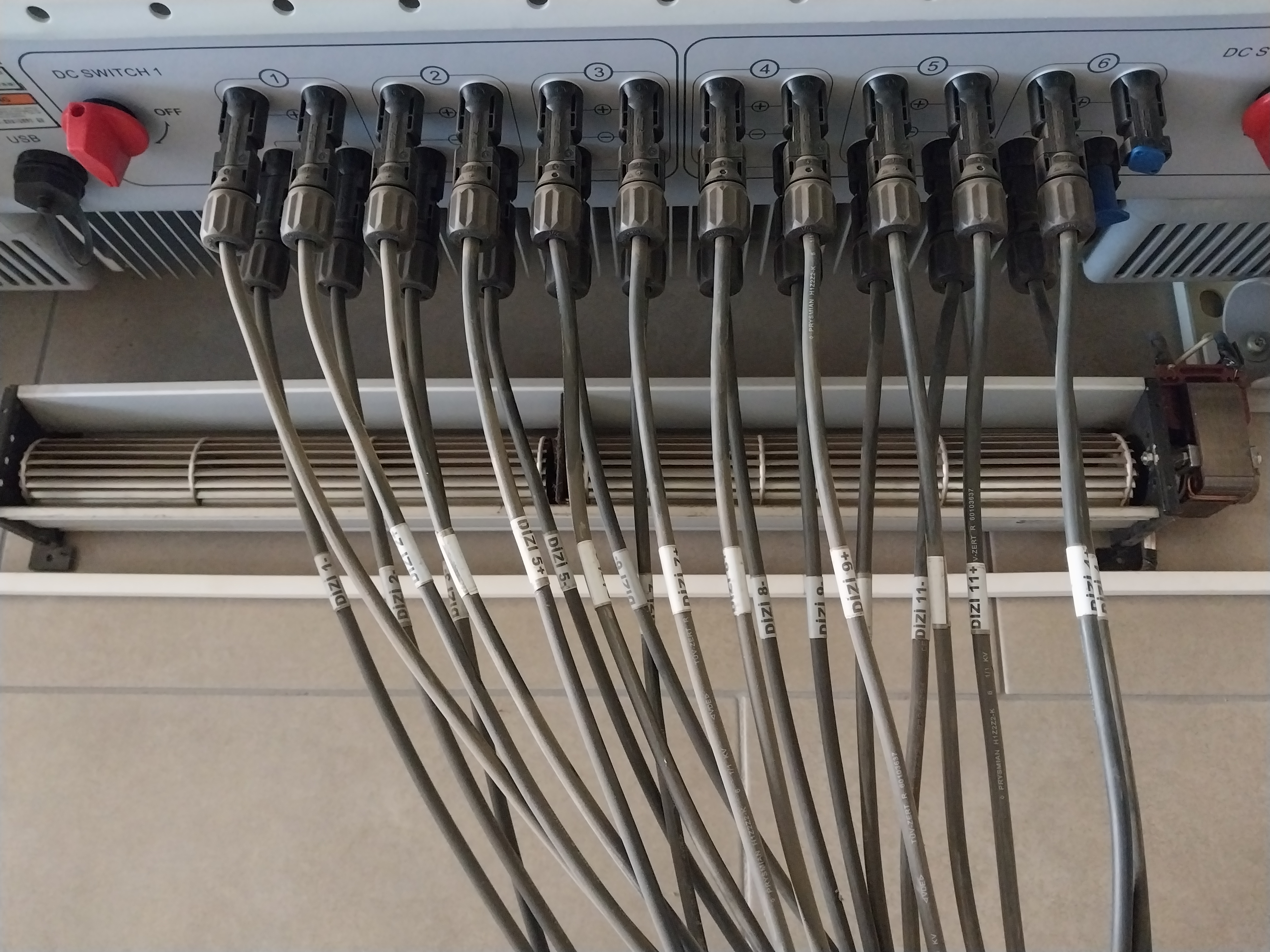 Yakından çekilmiş, DC SWITCH 1 etiketli DC dağıtım panosunda numaralandırılmış girişlere bağlı çoklu MC4 konnektörleri ve kabloları gösteren detay fotoğrafı. Kabloların üzerinde dizi numaralarını belirten beyaz etiketler bulunuyor, alt kısımda kablo kanalı ve zemin seramikleri görünüyor. GES DC tarafı bağlantı düzenini ve profesyonel montajı vurgulayan teknik bir görüntü.